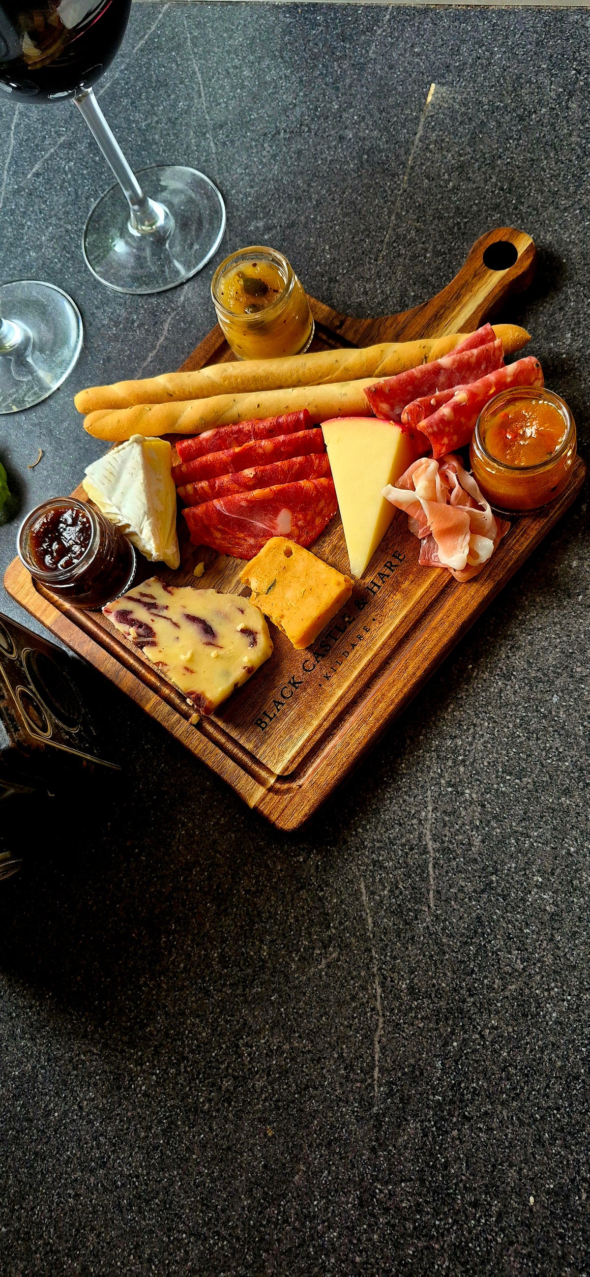 A charcuterie board with an assortment of cheeses, cured meats, breadsticks, and small jars of spreads, arranged on a wooden board over a dark textured surface with wine glasses partially visible.
