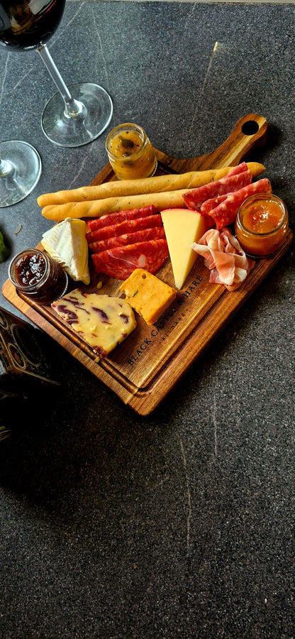 A charcuterie board with an assortment of cheeses, cured meats, breadsticks, and small jars of spreads, arranged on a wooden board over a dark textured surface with wine glasses partially visible.