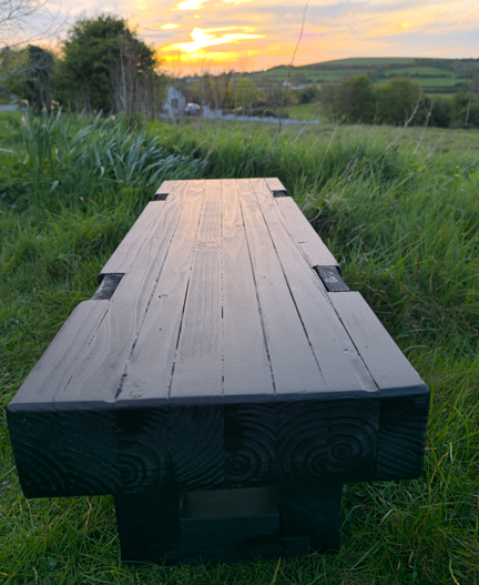 Black ash 2 seater wooden bench detail in a grassy field with a sunset in the background